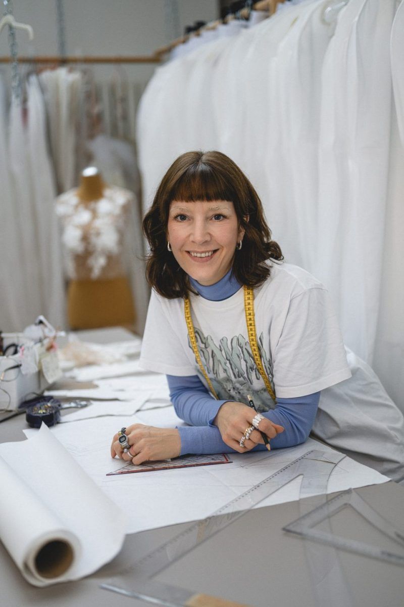 Beraterin im Brautmoden-Atelier in Köln mit Schnittmustern, umgeben von Brautkleidern und Maßband am Hals